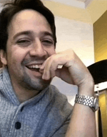 a man wearing a grey sweater and a silver watch is smiling with his hand on his chin .