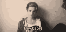 a young man in a hoodie is standing in front of a wall and says `` hey baby '' .