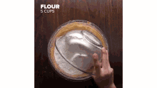 a person is adding flour to a glass bowl on a wooden table .