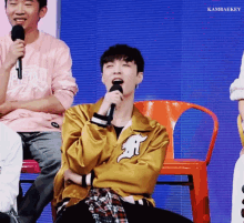 a man in a yellow jacket is singing into a microphone while sitting in a chair .