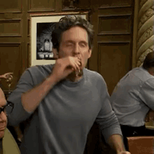a man in a grey shirt is drinking from a glass in a room .