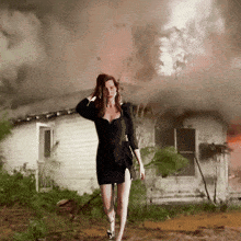a woman in a black dress is walking in front of a house that is on fire