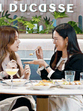 two women are sitting at a table with plates of food and drinks in front of a sign that says ducasse