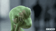 a green lizard is covering its face with its hands and the word geico is on the bottom .