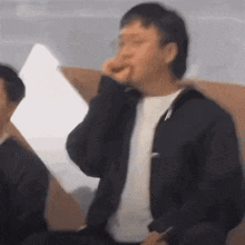 a man in a black jacket is sitting on a couch covering his nose with his hand .