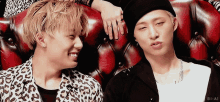 two men are sitting next to each other on a red couch and one of them is wearing a beanie