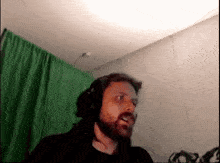 a man with a beard is wearing headphones in a room with a green curtain .
