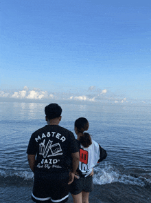 a man wearing a master said shirt stands next to a woman looking out over the ocean