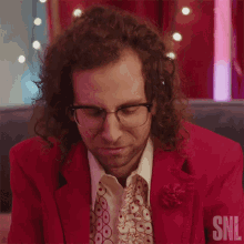 a man with curly hair and glasses is wearing a red suit and tie with the snl logo in the corner