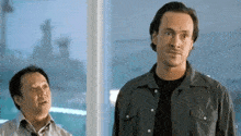 two men are standing next to each other in front of a window and one of them is taller than the other