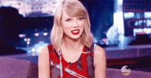 taylor swift is sitting on a couch in a red dress with red lipstick on her lips .