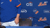 a man in a blue mets jersey sits at a table with microphones in front of a wall that says citi field