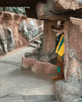 a cartoon character in a yellow and green outfit stands under a rock