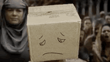 a woman is holding a cardboard box with a sad face drawn on it in front of a crowd of people .
