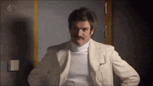 a man in a white suit and turtleneck is standing in front of a door with his hands on his hips .