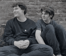 two young men are sitting next to each other on a sidewalk in front of a brick wall .