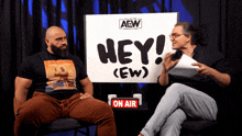 two men sitting in front of a sign that says hey