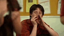 a man in a red shirt is covering his nose with his hands while sitting at a table .