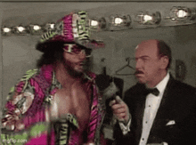 a man in a tuxedo is talking to a wrestler in a colorful hat and sunglasses .