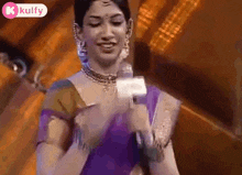 a woman in a purple saree is holding a microphone and talking into it .