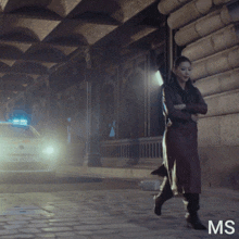 a woman in a trench coat is walking down a street next to a police car