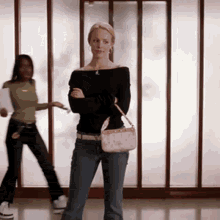 a woman is standing with her arms crossed and holding a pink purse while another woman dances in the background .