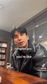 a man in a black leather jacket is sitting at a table with a caption that says hongdae guy you 're like