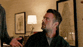 two men are talking in a living room with a lamp in the background .