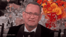 a man in a tuxedo and bow tie is sitting at a table with a vase of flowers in the background .