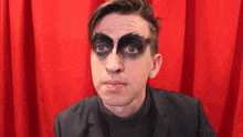 a man with black makeup on his face looks at the camera in front of a red curtain