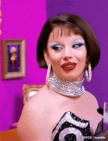 a drag queen wearing a choker and earrings is standing in front of a pink wall .