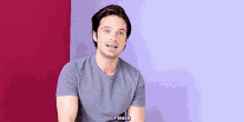 a man in a grey t-shirt is sitting in front of a purple wall and talking .
