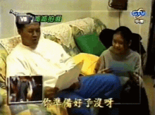 a man sits on a couch reading a book while a woman sits next to him with chinese writing on the screen behind them