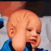 a baby in a blue shirt is making a funny face with his hand on his forehead .