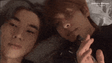 two men are laying next to each other on a bed and one of them is making a heart with his finger .