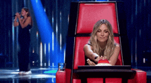 a woman in a red chair is pressing a button while another woman sings behind her