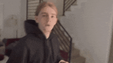 a young man in a black hoodie is standing in a living room next to stairs .