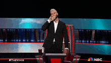 a man in a suit is covering his mouth while sitting in a chair on a stage with a nbc logo in the background