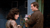 a man and a woman are standing in front of a blue door and talking about flowers