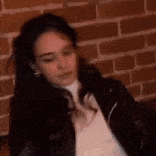 a blurry picture of a woman sitting in front of a brick wall wearing a leather jacket .