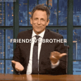 a man in a suit and tie is sitting at a table with his arms outstretched and the words friends vs brother behind him .