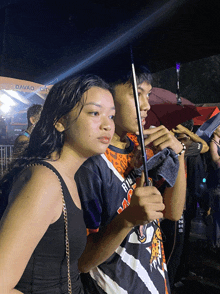 a man holding an umbrella stands next to a woman wearing a shirt that says ' rm '