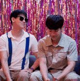 two men wearing sunglasses are sitting on a couch in front of a pink and purple curtain