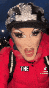 a drag queen wearing a helmet and a red jacket says the on her face .