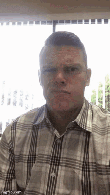 a man in a plaid shirt is making a funny face while sitting in front of a window .