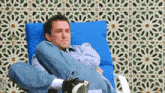 a man is sitting in a blue chair with his legs crossed and a patterned wall behind him .