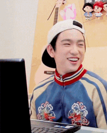 a young man wearing a baseball cap and a jacket smiles in front of a laptop