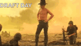 a shirtless man in a cowboy hat is standing in a field with soldiers in the background .