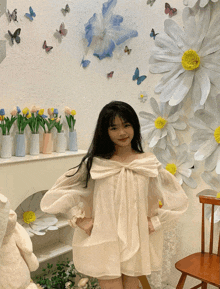 a little girl in a white dress stands in front of a wall of paper daisies and butterflies