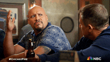 two men are sitting at a table with a bottle of beer in front of them and a nbc logo in the corner